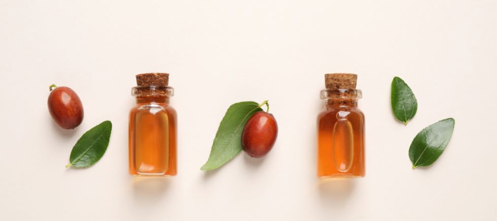 Diferentes botes de aceite de jojoba con hojas y semillas sobre fondo blanco roto.