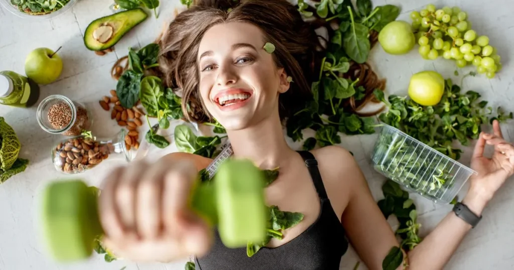 Mujer sonriendo levantando una pesa verde sobre fondo de verduras y hortalizas o "beauty food".