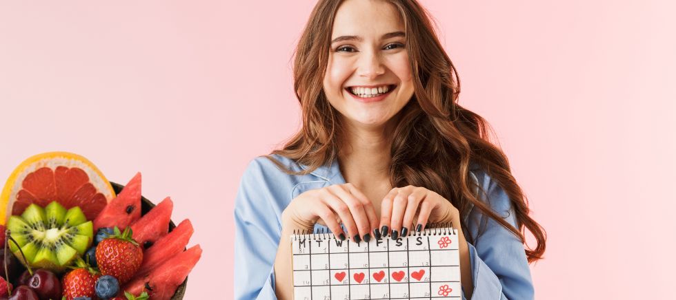 Joven con un calendario de frutas y verduras de temporada en la mano y una cesta de fruta al costado.